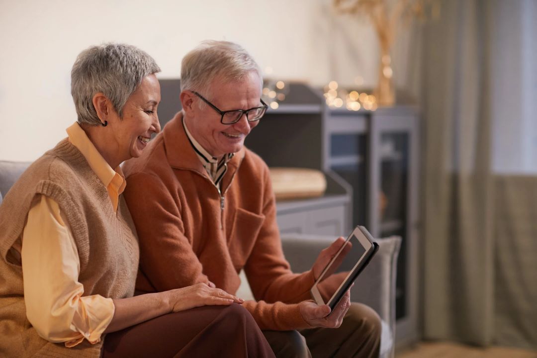 Two older adults sitting together, looking down at a tablet and smiling.